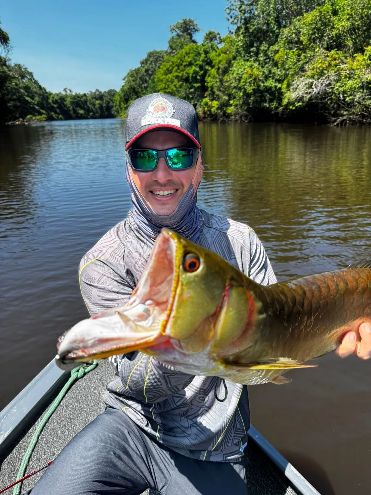 Man smiling on a boat holding a large fish with a river and green trees in the background.