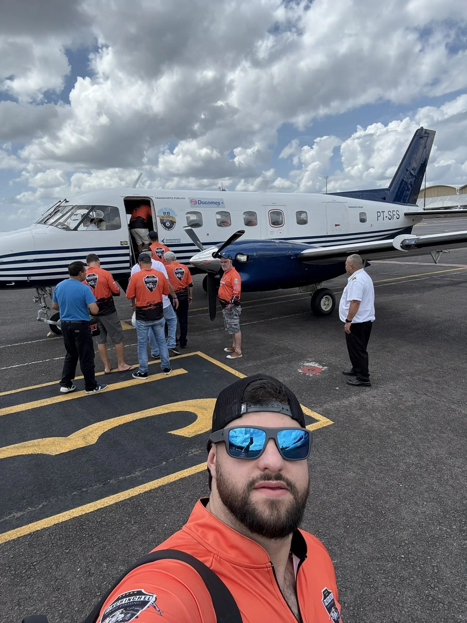 A man with sunglasses and a red jacket taking a selfie in front of a small aircraft on the tarmac, with several people entering the plane, and a partly cloudy sky overhead.