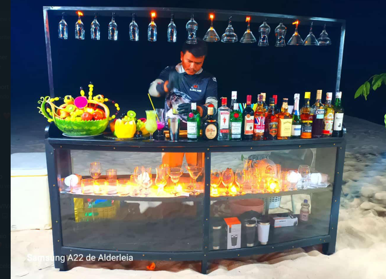 A bartender preparing drinks at a beachside bar with alcohol bottles, glassware, a fruit bowl, and illuminated candles, under hanging glasses.