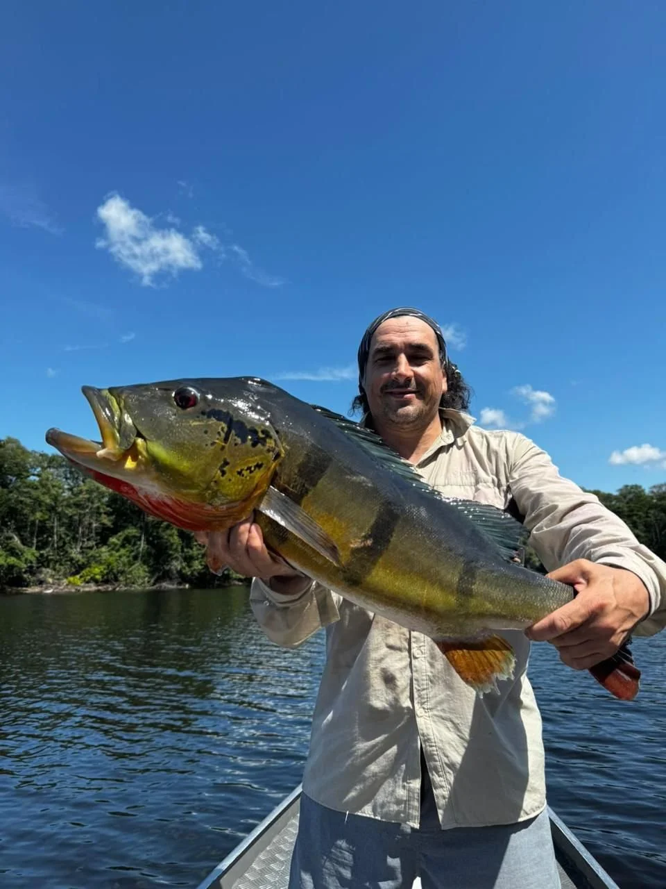 Man holding a large fish on a boat in a river, with trees and blue sky in the background.