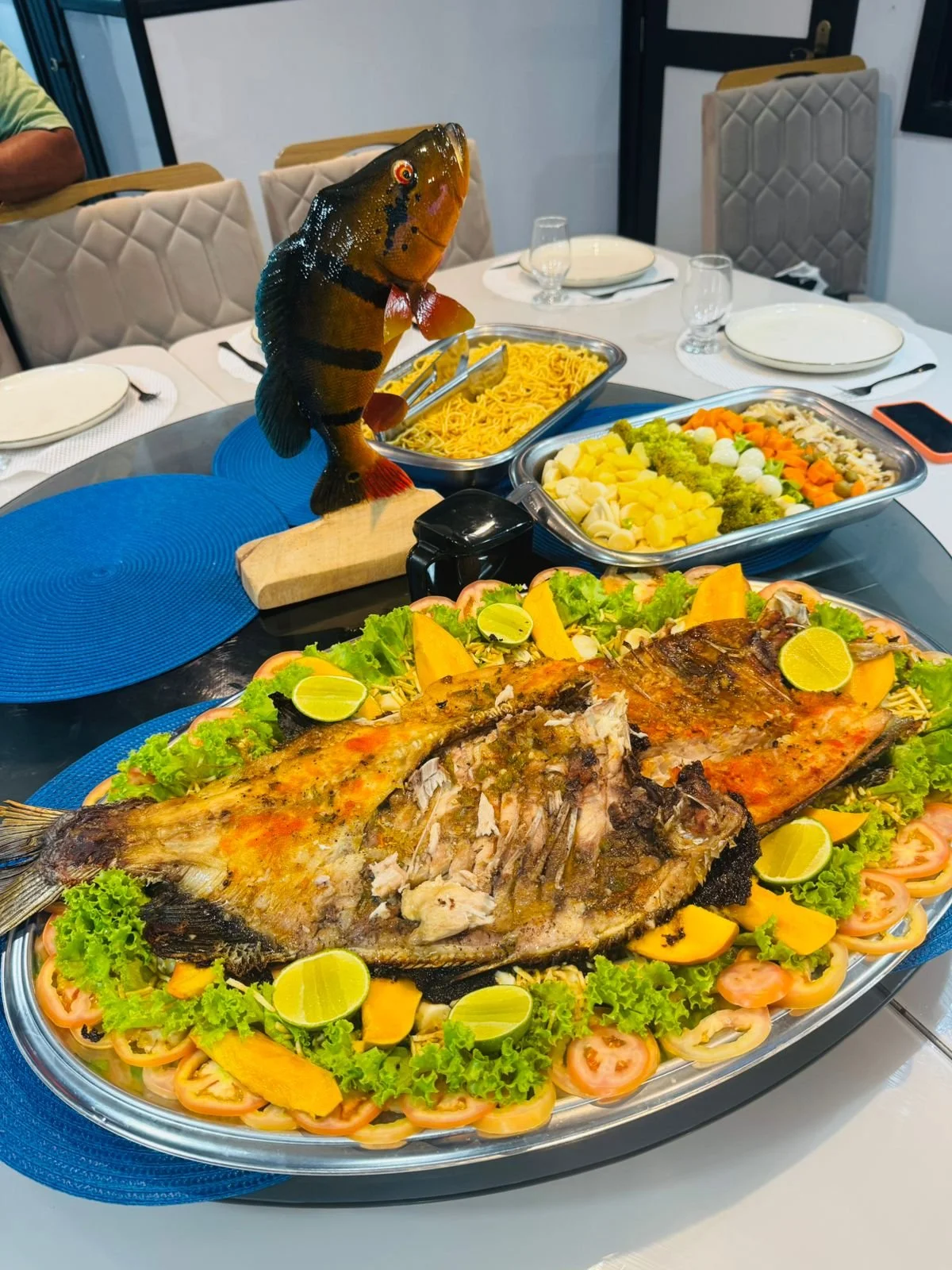 A large roasted fish garnished with lime slices, surrounded by lettuce, tomatoes, and sliced yellow vegetables on a silver platter. In the background, there are bowls of pasta, boiled potatoes with vegetables, and a fish sculpture in a dining setting