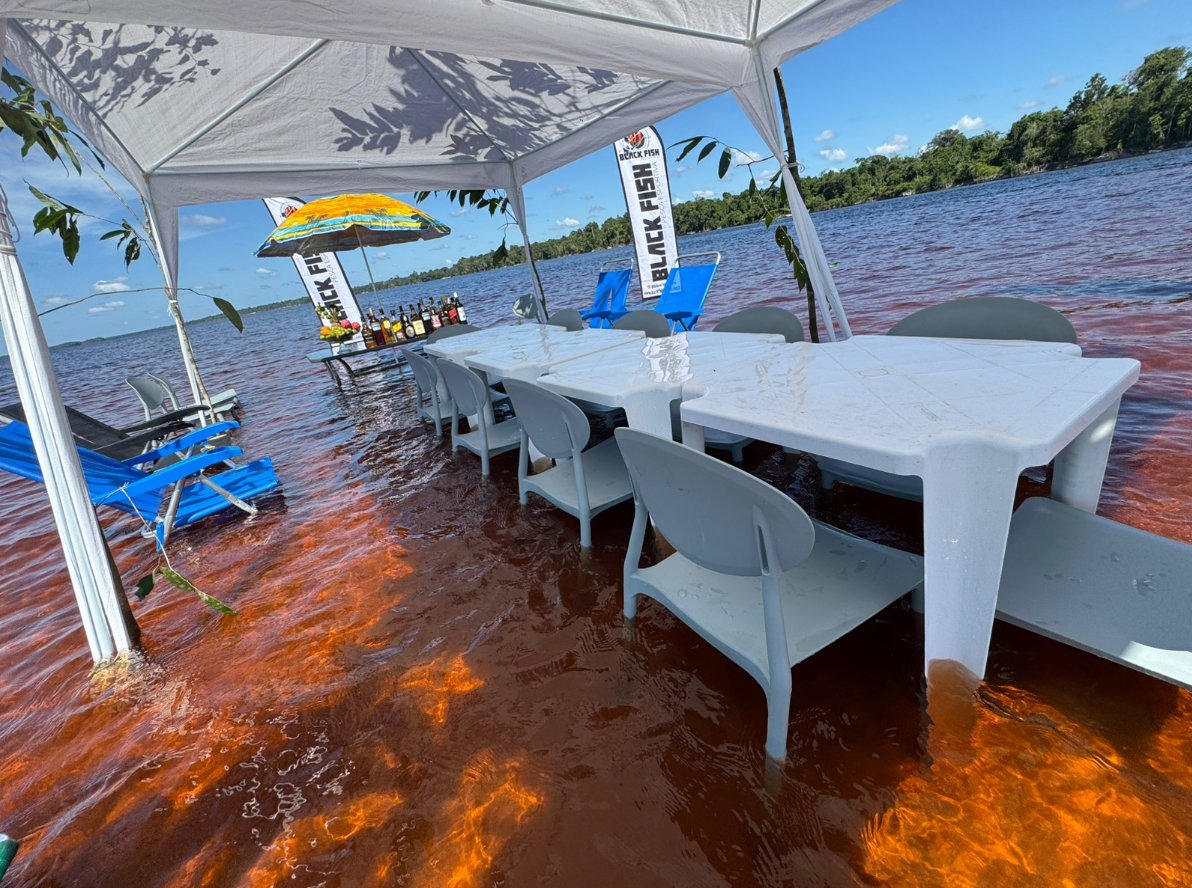 Outdoor dining area with a table and chairs under a tent, set up on a river with water reaching the chairs. There are blue lounge chairs, a table with drinks, and a colorful umbrella nearby. Flags and trees are visible in the background.