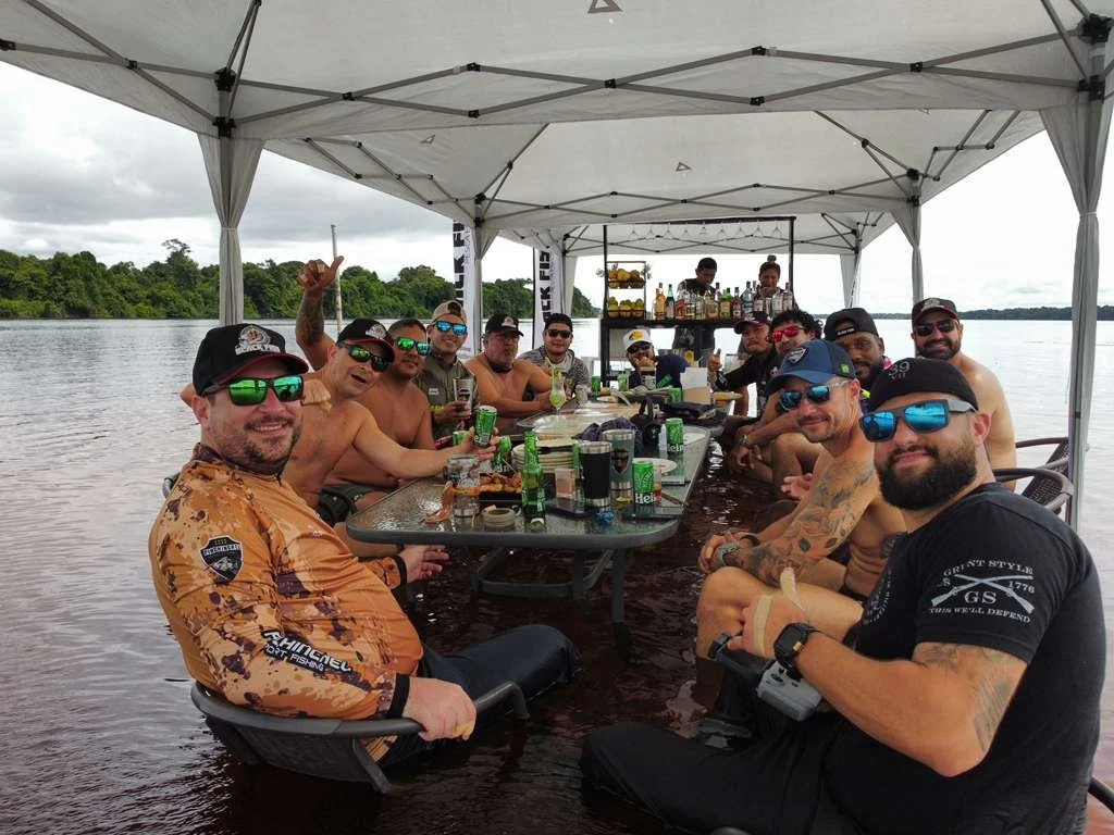 A group of men sitting around a table on a boat by a river, enjoying drinks and food under a canopy