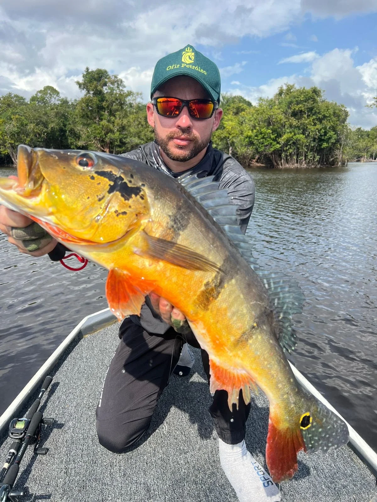 Man holding a large fish on a boat in a river with trees and cloudy sky in the background.