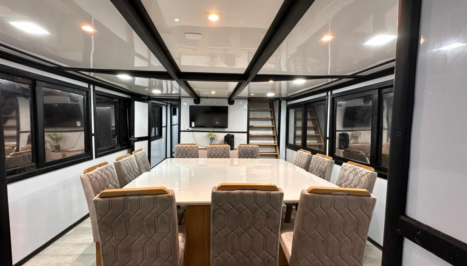A modern conference room with a large white table surrounded by beige upholstered chairs, black-framed glass walls, a flat-screen TV on the back wall, and wooden stairs leading to an upper level.