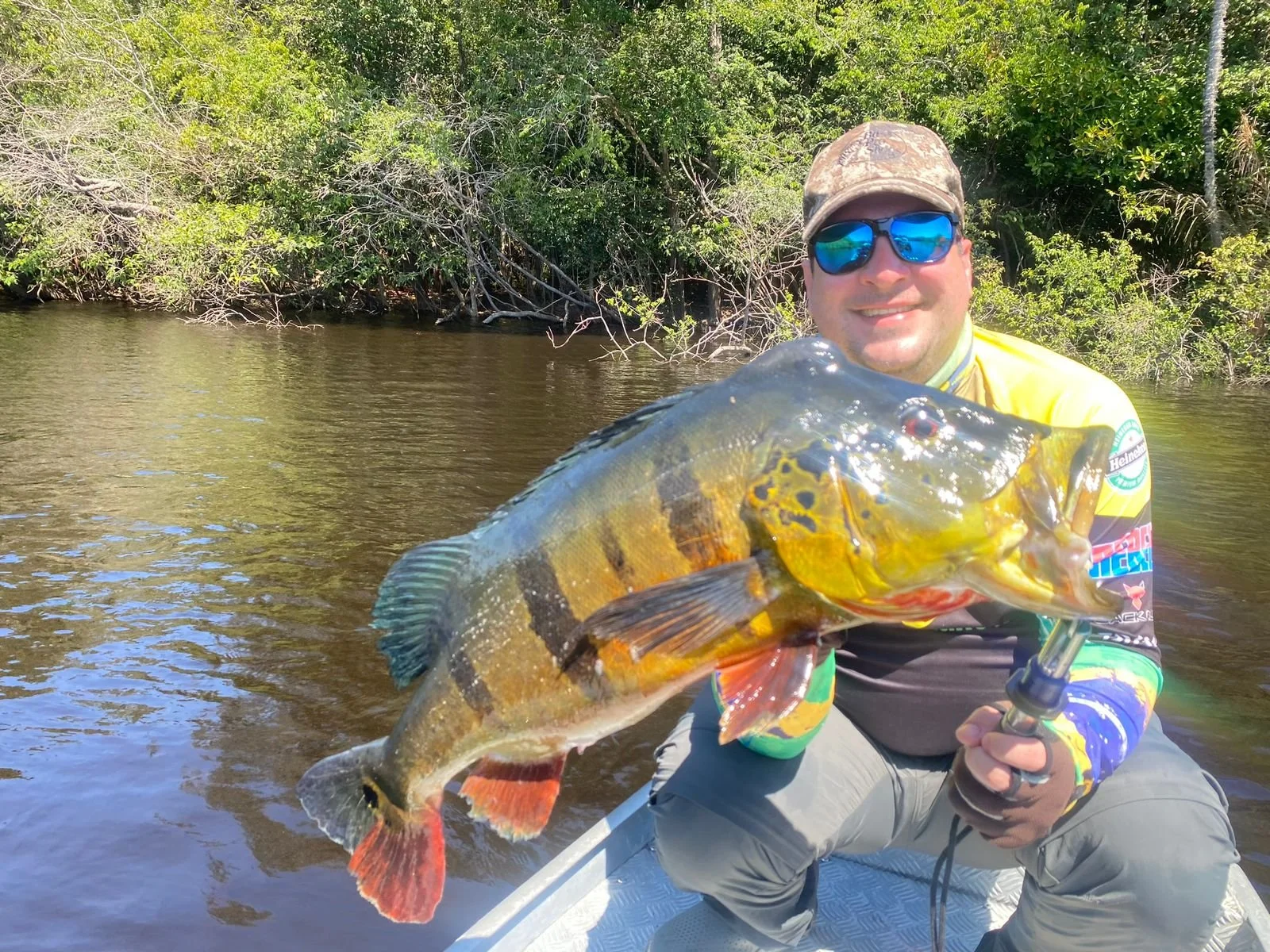A man wearing sunglasses, a camouflage cap, and a yellow, black, and blue fishing jersey is holding a large colorful fish with red, yellow, and black markings, on a boat or small watercraft on a river with dense green trees in the background.
