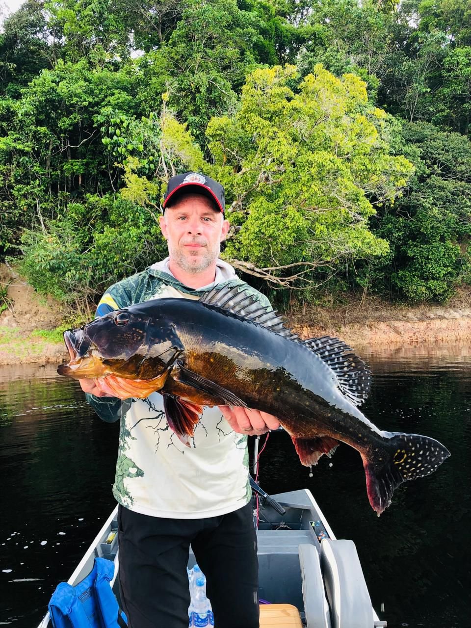 Man holding a large fish on a boat with a background of lush greenery and trees by a waterbody.