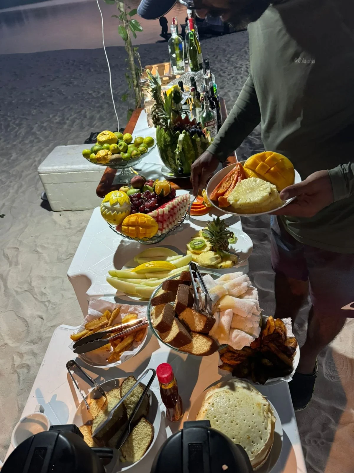 Beachside buffet table with assorted fruits, cakes, and beverages, man selecting food.