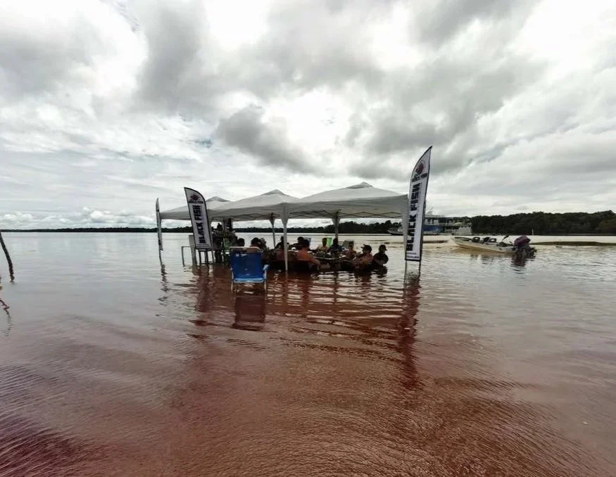 Group of people under a white canopy tent on a flooded body of water with grey cloudy sky, flags with 'Black Fish' text, and a boat in the background.