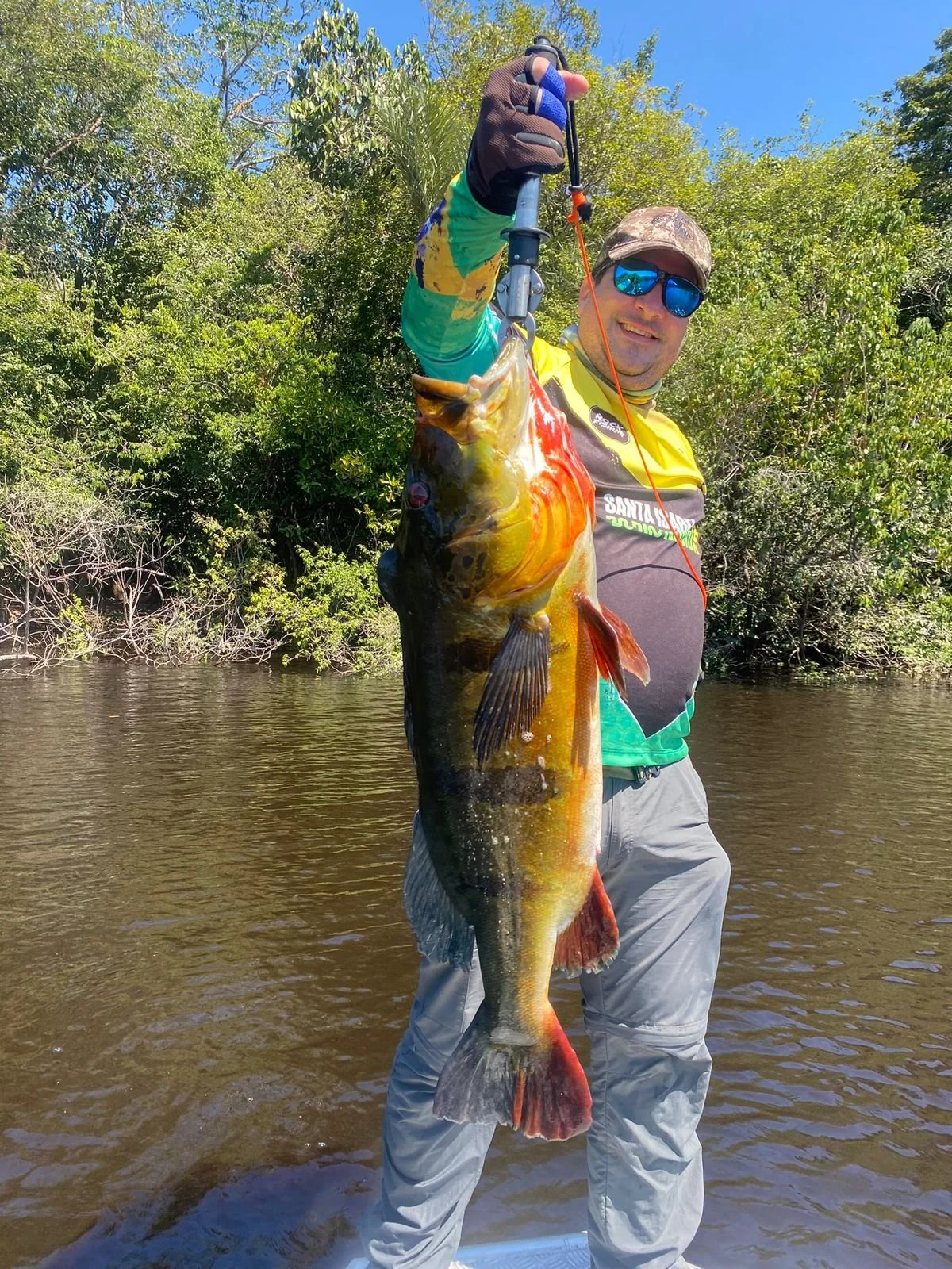 Man in sunglasses and fishing gear holding a large fish with a fishing plier, standing in a river with trees and blue sky in background.