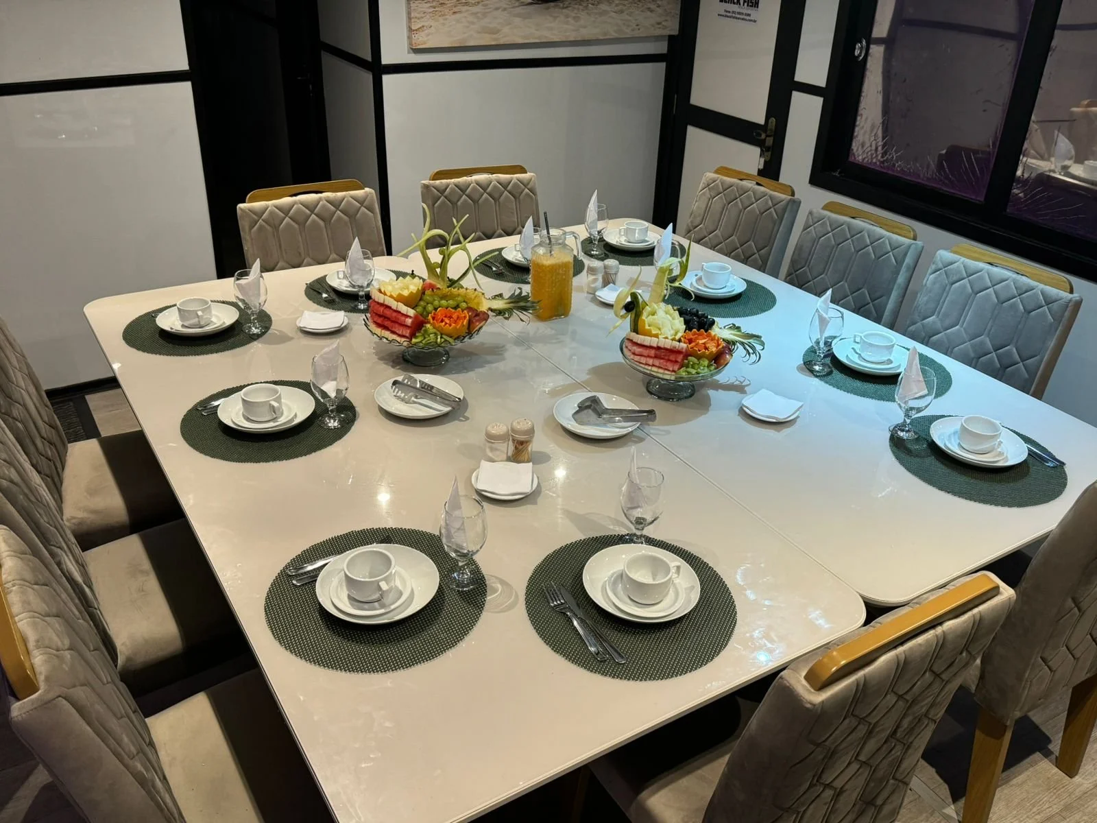 A dining table set for eight with green placemats, white cups and saucers, and silverware. There are two large bowls of fruit in the center, with fruit and a glass of orange juice nearby.