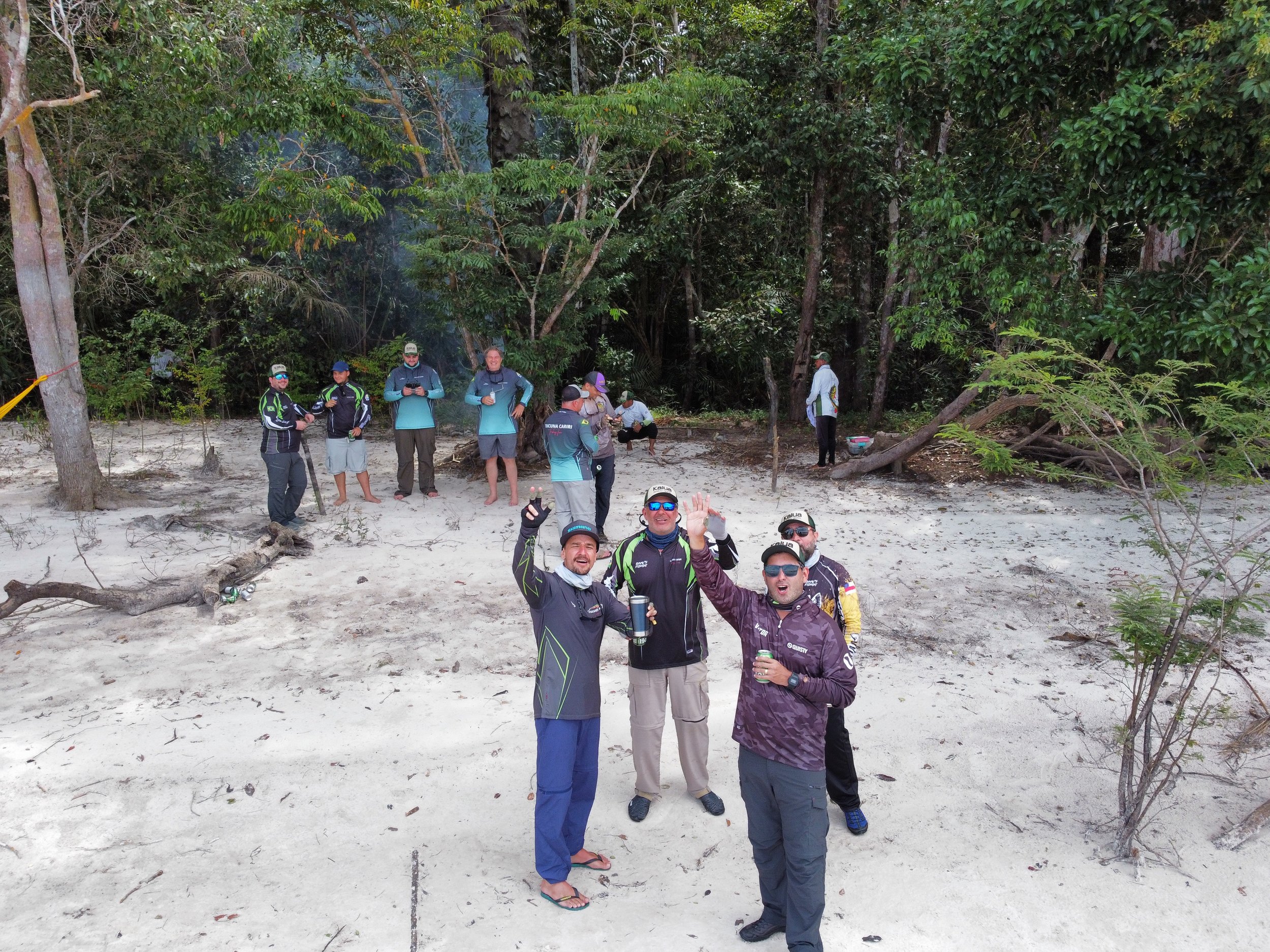 Group of people on a beach, some are standing and others are sitting or walking, with trees and foliage in the background, celebrating and smiling.
