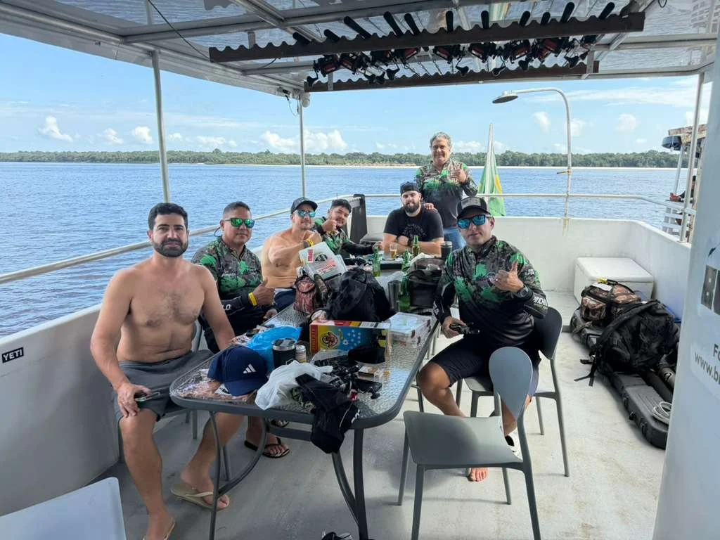 Group of six men sitting and standing on a boat deck, with fishing gear and snacks, overlooking a body of water under a partly cloudy sky.