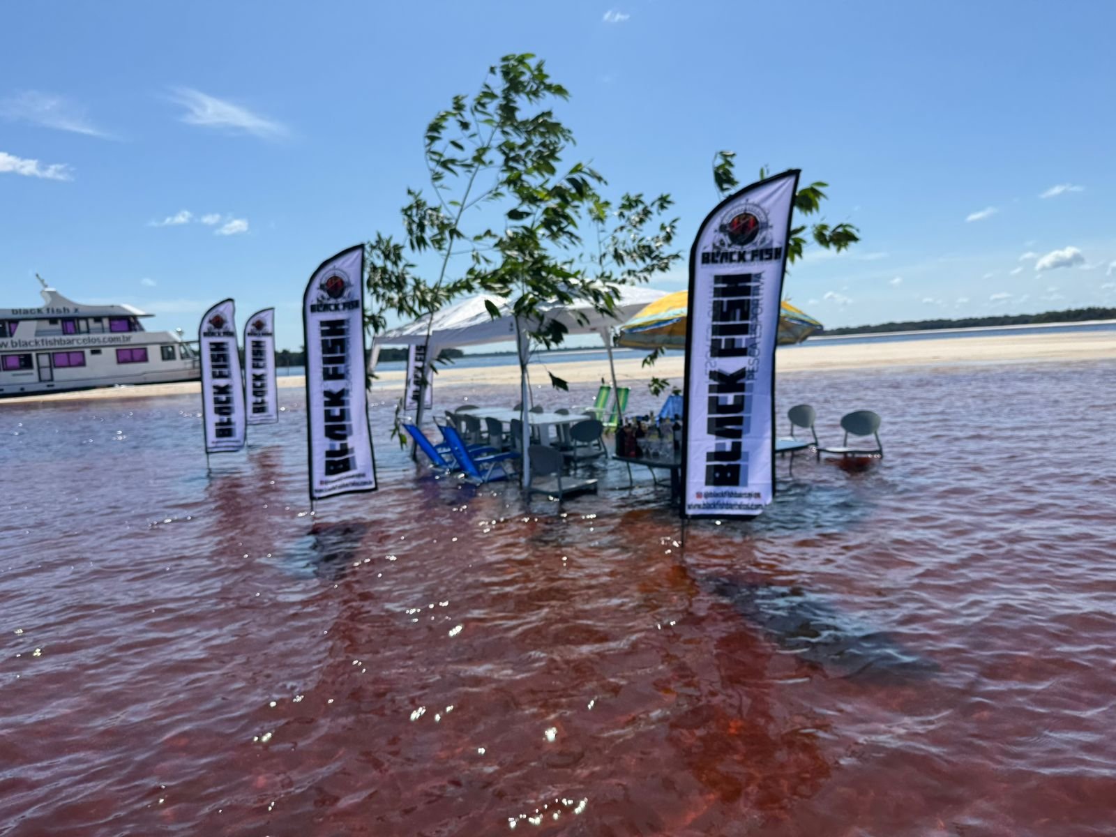 A floating pavilion on reddish water with several black flags labeled 'Black Fish' and a tree with green leaves in the foreground. There are chairs, tables, and umbrellas on the pavilion, and a boat is docked nearby.