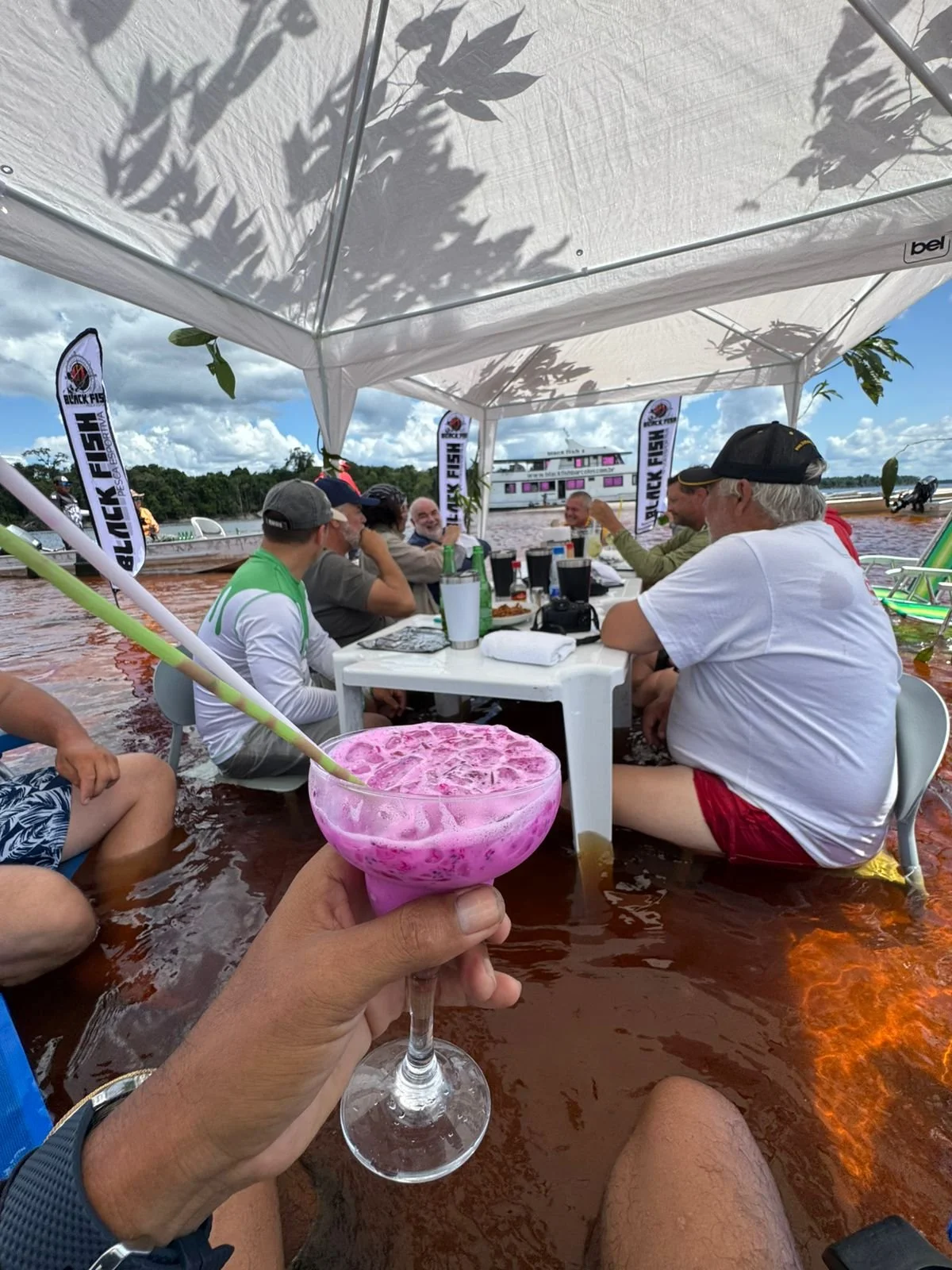 People sitting in a boat on a river under a canopy, with a man holding a pink tropical cocktail with a straw in the foreground, enjoying a social gathering.
