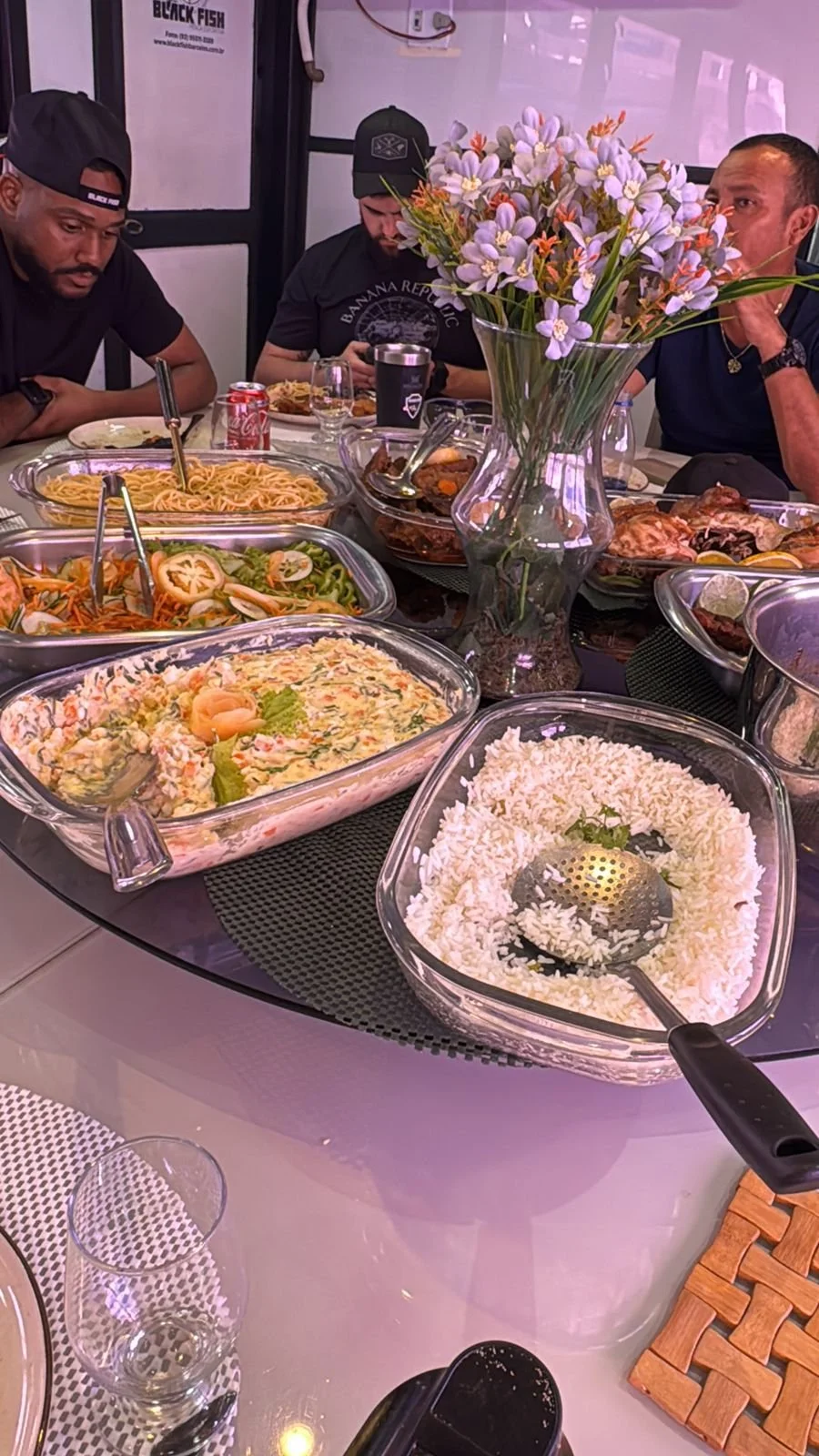 A dinner table with various dishes including rice, salad, pasta, and a large vase of purple flowers, with three men sitting around it.