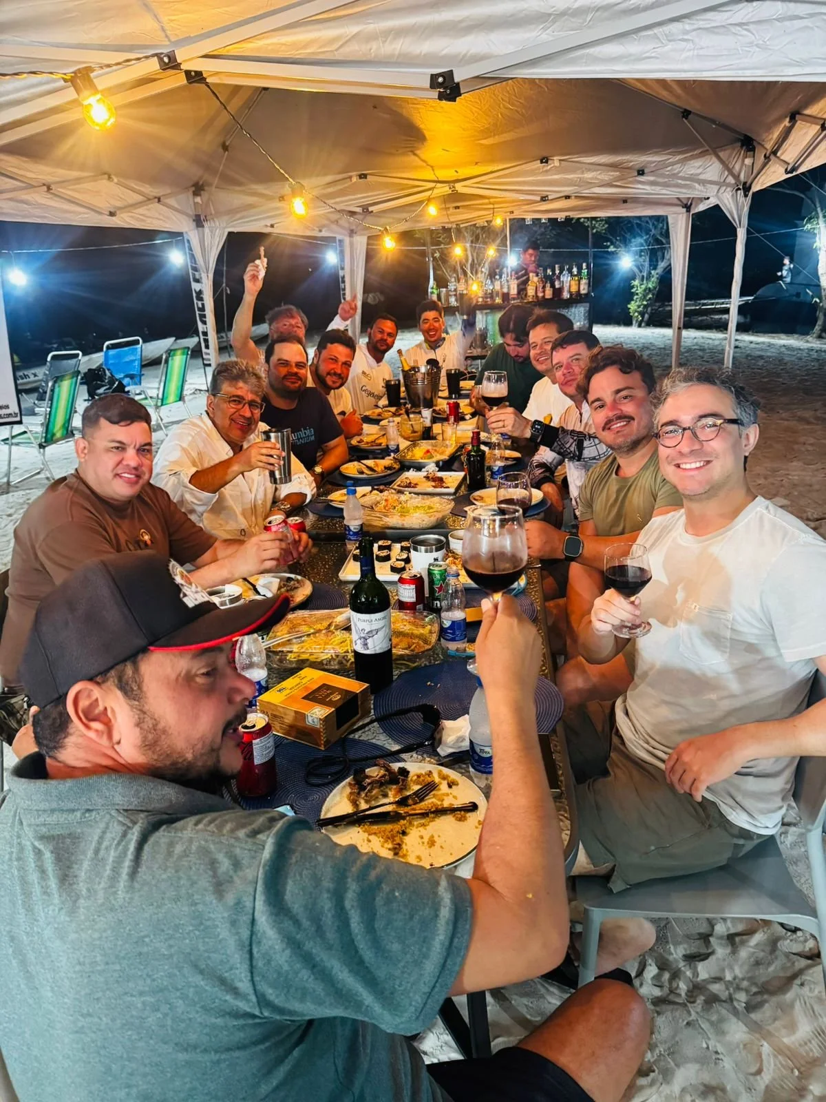 Group of people gathered around a long table under a tent at night, enjoying food and drinks, with string lights overhead and drinks like wine and beer visible on the table.