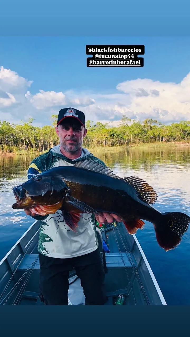 A man in a boat holding a large dark-colored fish with a scenic background of trees and a blue sky with clouds.