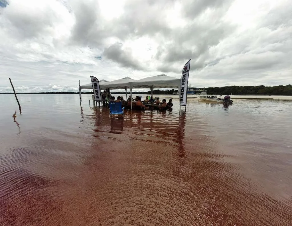 A group of people sitting under a white canopy on a beach with red sand, near the water, with flags on either side of the canopy, overcast skies, and boats on the water in the background.
