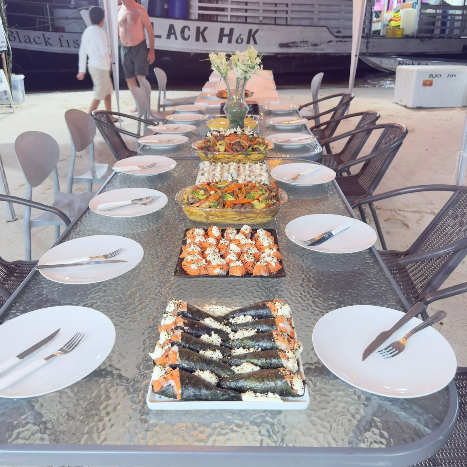 A long glass outdoor dining table set with white plates, silverware, and empty glasses, with various sushi dishes, vegetable salads, and a flower vase in the center. The background features a boat and sandy ground.