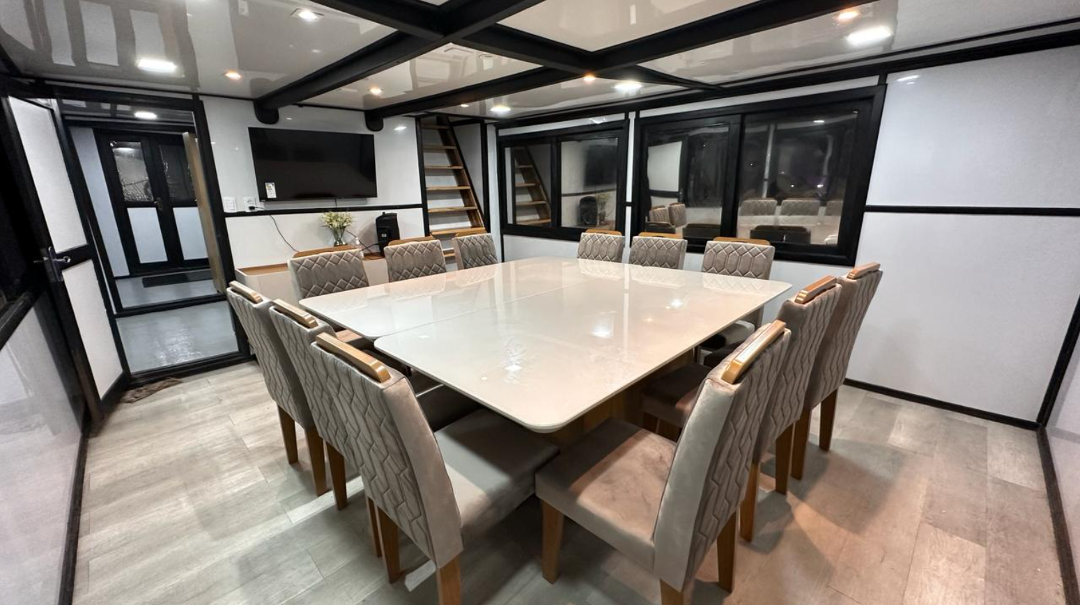 Conference room with a large rectangular table and 12 upholstered chairs, a wall-mounted TV, a small plant, and large windows.