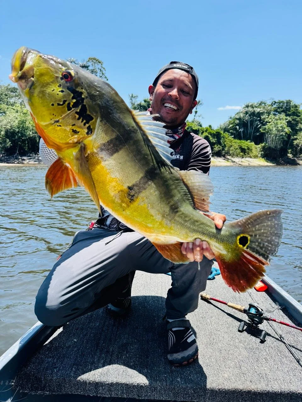 A man in fishing gear holding a large yellow and green fish with black markings, on a boat in a river with trees and blue sky in the background.