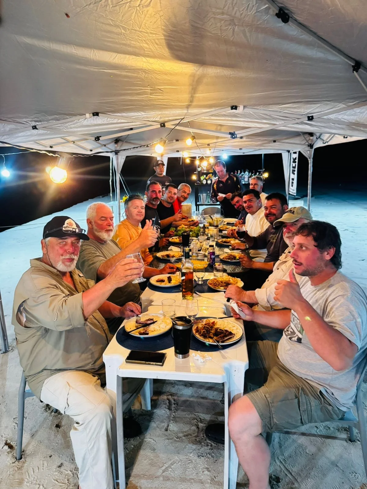 A group of men enjoying a dinner party under a white canopy at night, with food and drinks on the table.