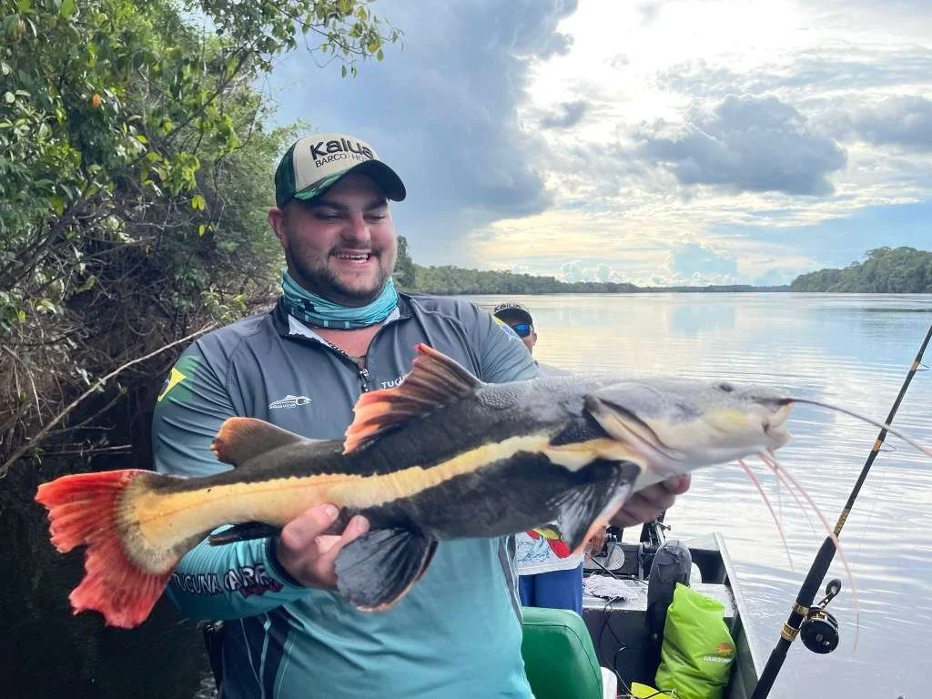 A man smiling and holding a large catfish he caught while fishing by a river. He is wearing a cap, a fishing shirt, and a neck gaiter, with a fishing rod and gear visible in the background.