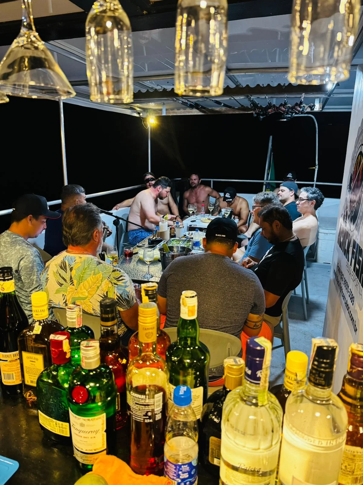 A group of men sitting around a table on a rooftop at night, some shirtless, with bottles of alcohol and glasses, engaged in conversation.