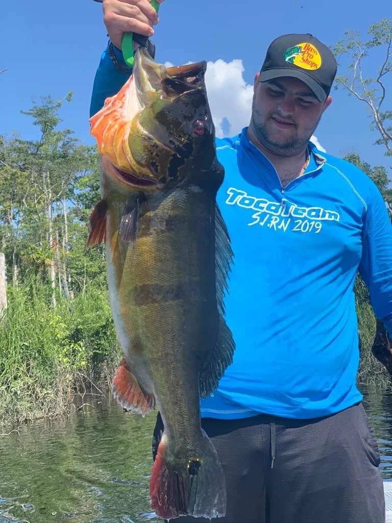 Man holding a large bass fish he caught, outdoors near water and trees, wearing a blue Tacklebox shirt and a gray Cap with a Bass Pro Shops logo.