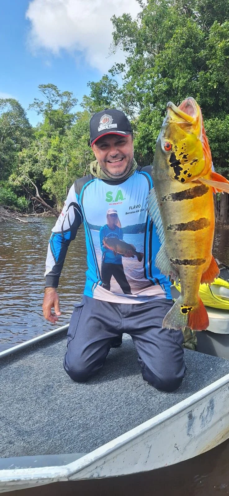 Man on a boat holding a large, colorful fish with green trees and a blue sky in the background.
