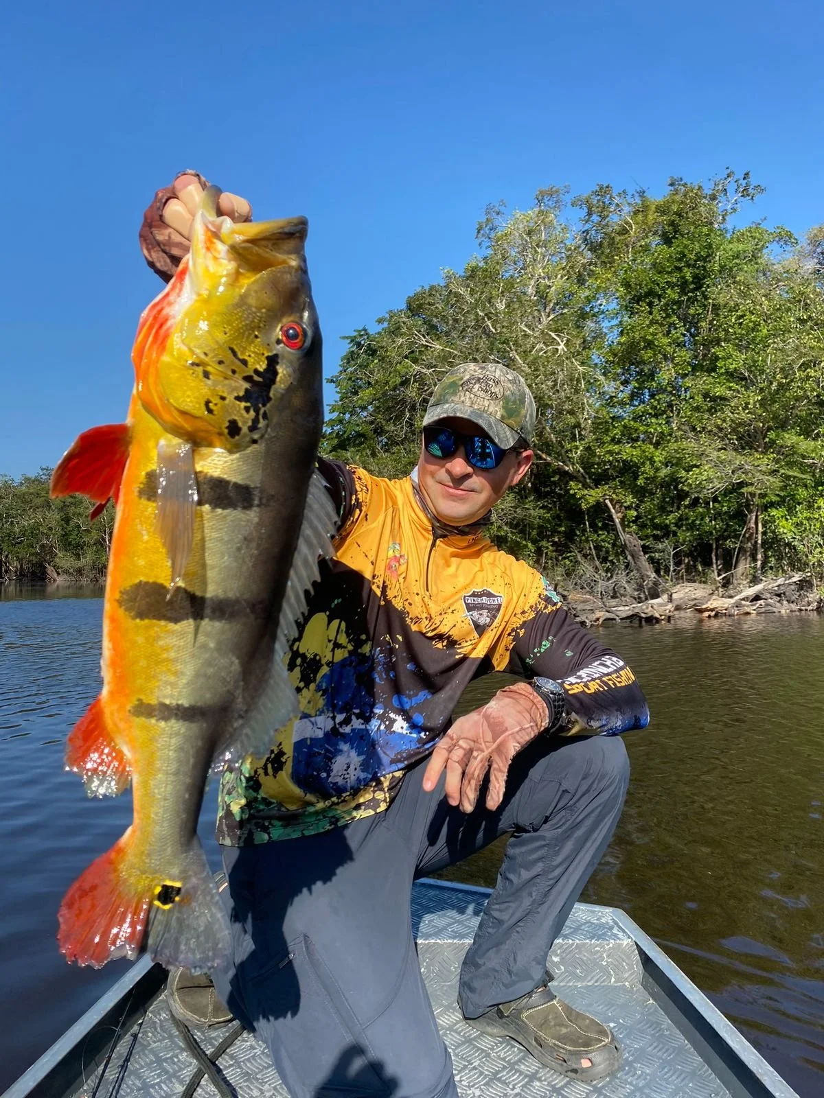 Man wearing sunglasses and a camouflage hat kneeling on a boat, holding a large colorful fish with a yellow, black, and orange pattern, against a backdrop of water and green trees under a clear blue sky.