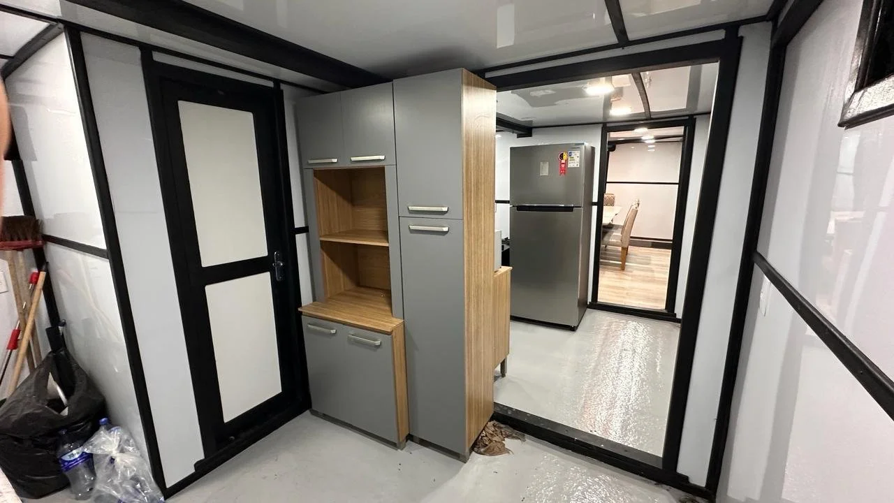 Interior of a small modern kitchen with gray cabinets, wooden open shelves, and a silver refrigerator, viewed through a black and white framed doorway with a reflective floor.