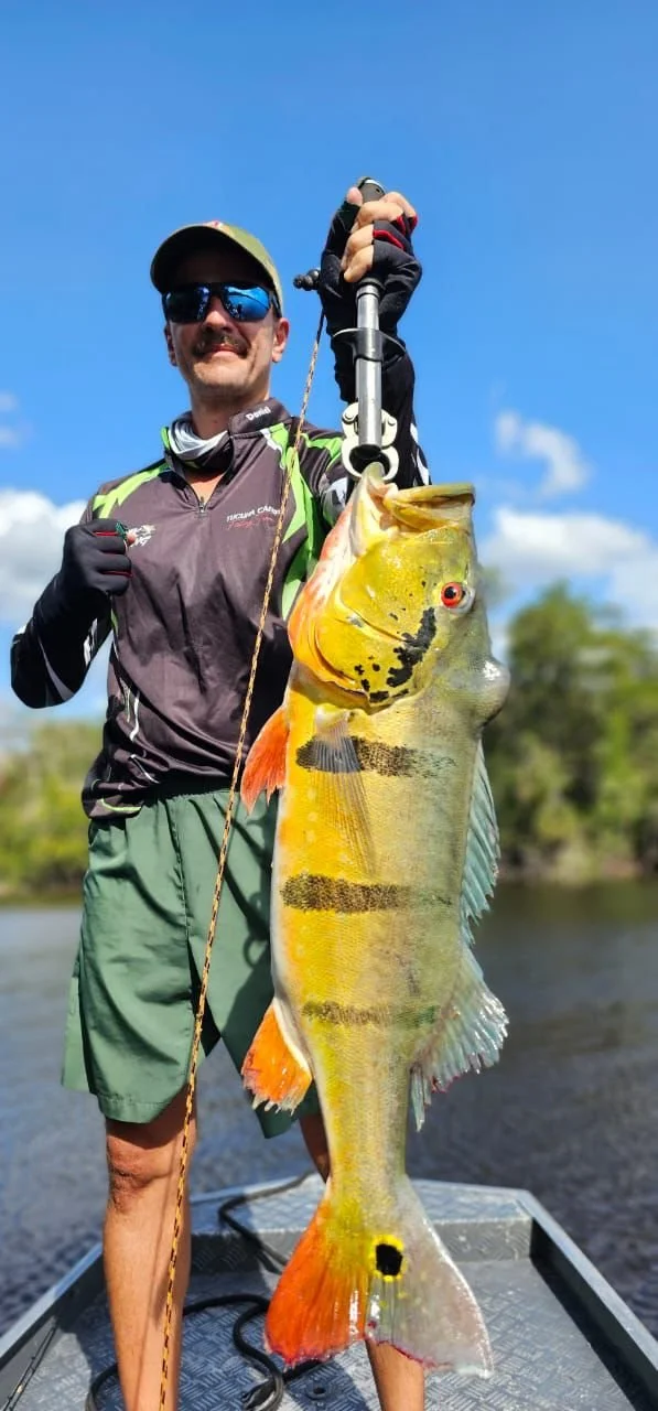 Man in sunglasses and cap holding a large fish with a measuring device, standing on a boat with water and trees in the background.