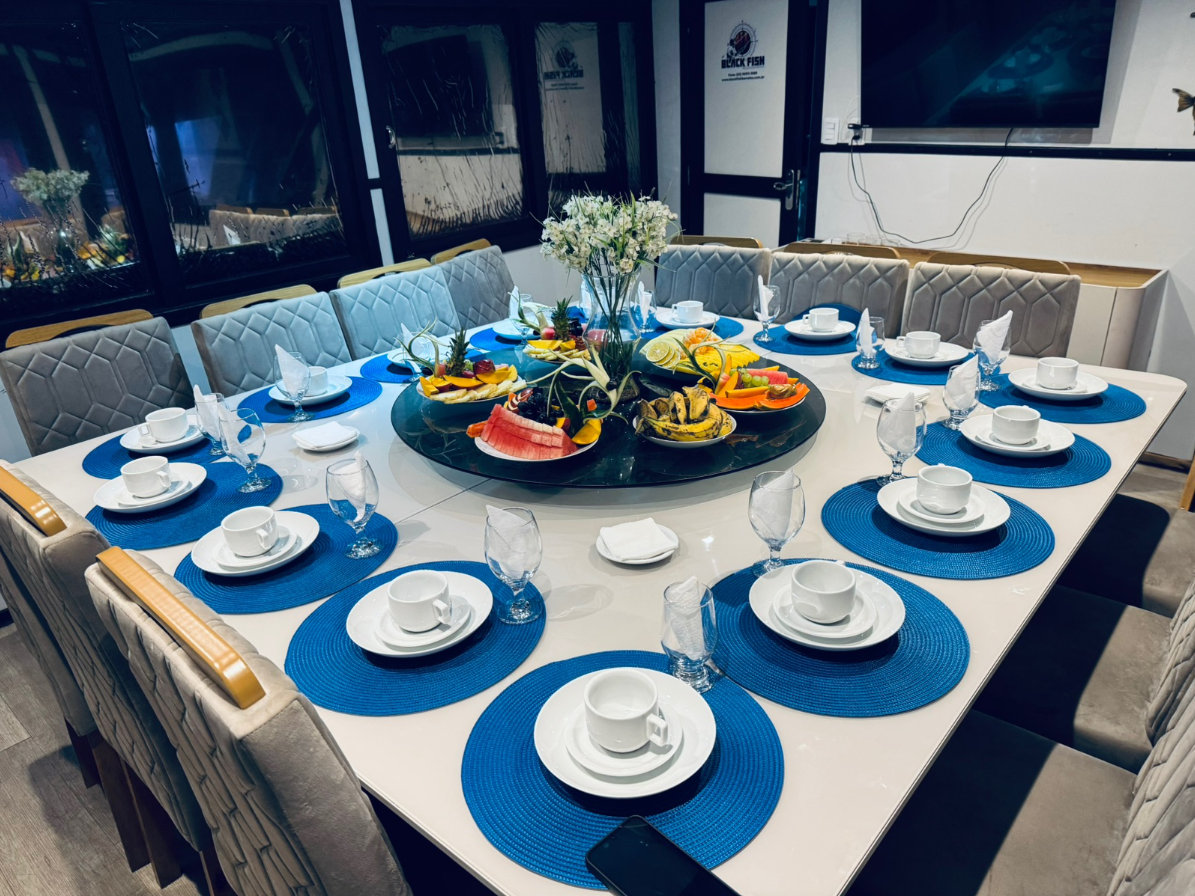 A round table set for a meal with white cups, plates, and glasses on blue placemats, with a floral centerpiece and fruit on the table, in a room with chairs and a television on the wall.