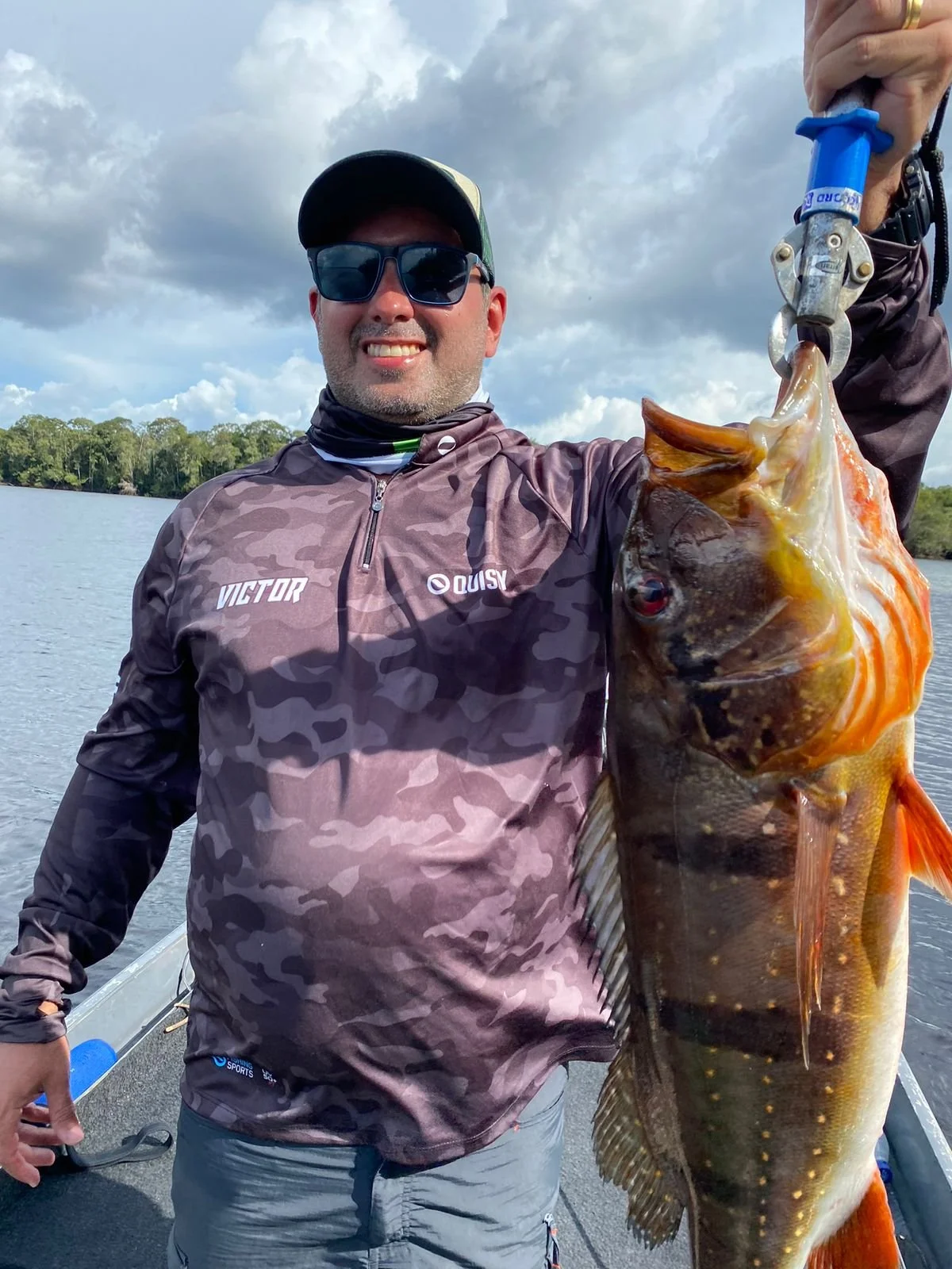 Man smiling while holding a large fish with a fish grip on a boat on a lake, with trees and cloudy sky in the background.