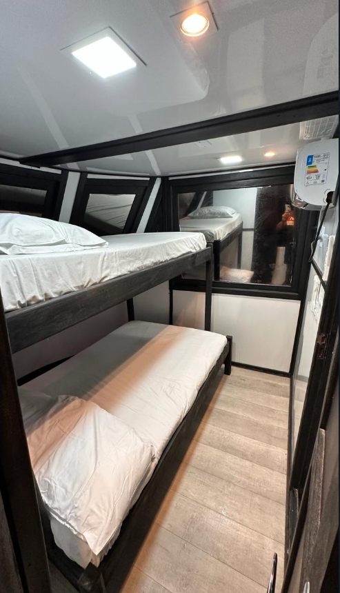 Inside a compact room with black loft beds, white bedding, and a window with a view outside; ceiling lights and a water heater are visible.
