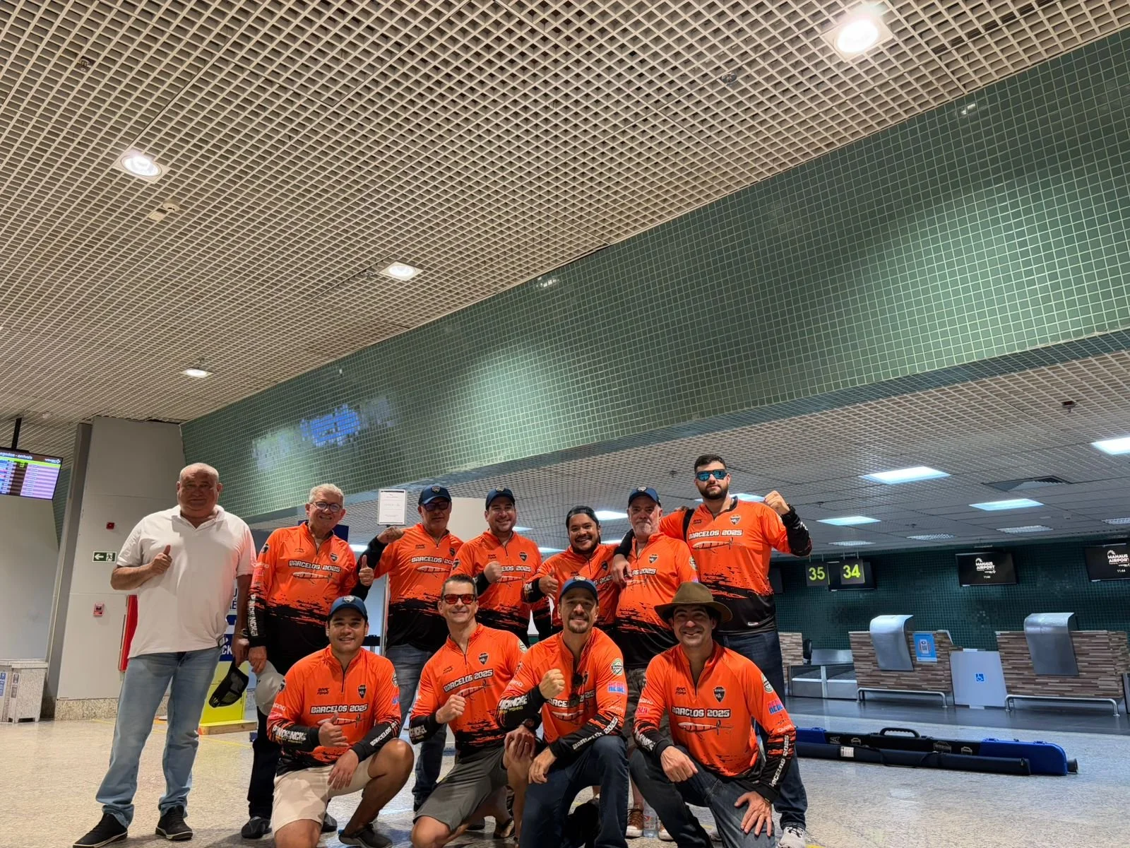 Group of ten men at an airport, some wearing orange jerseys with race or team logos, posing for a photo, with two men giving thumbs up, one wearing sunglasses and a hat, in front of check-in counters.