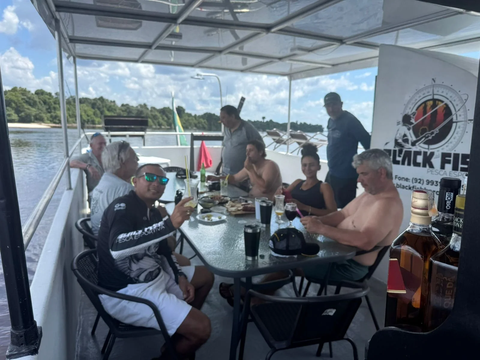 Group of people enjoying drinks and food on a boat deck with a river and trees in the background, during daytime.