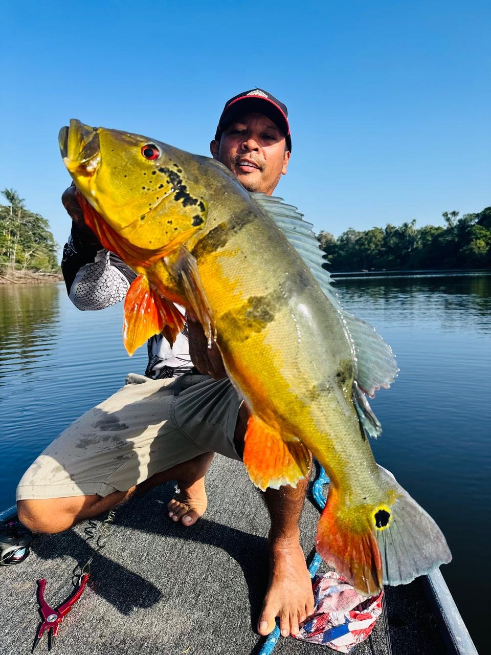 Man holding a large fish on a boat in a river with trees in the background.