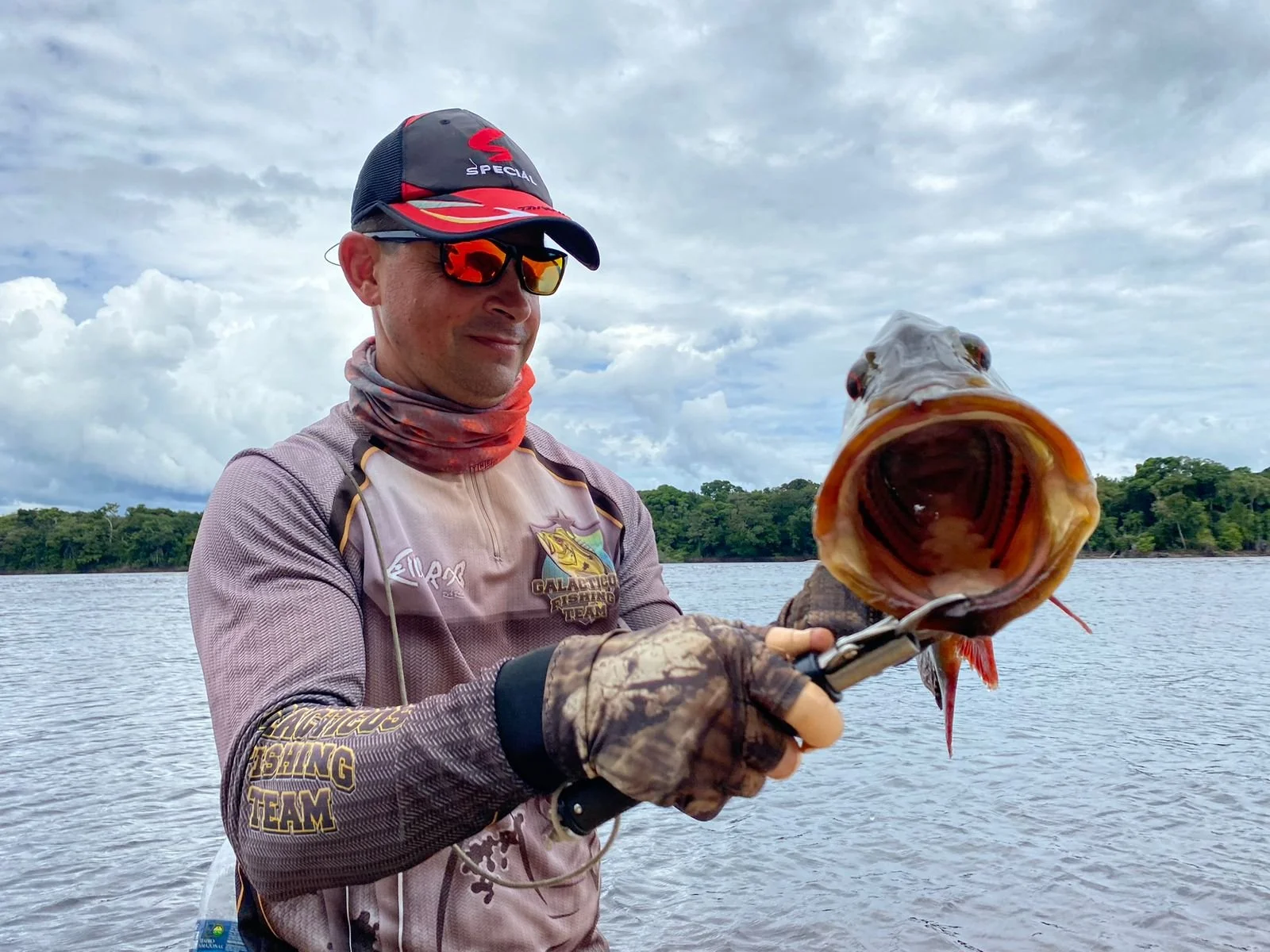 A man wearing a fishing cap, sunglasses, and fishing gear holding a large fish with an open mouth near a body of water, with a cloudy sky and trees in the background.