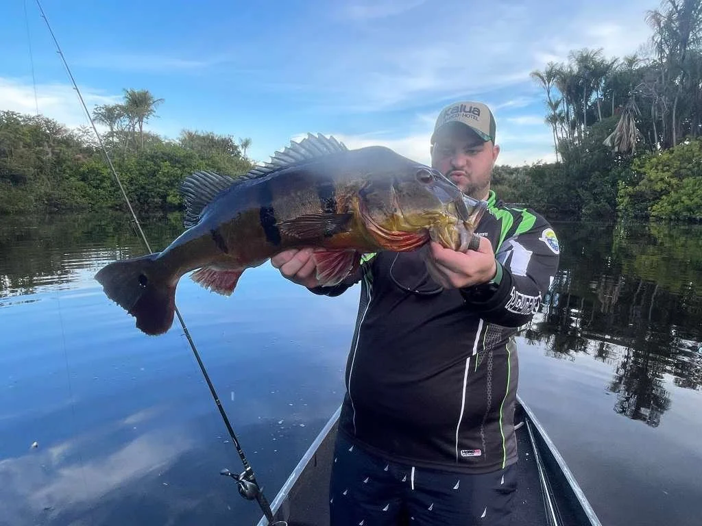 A man holding a large fish on a boat in a river surrounded by trees.
