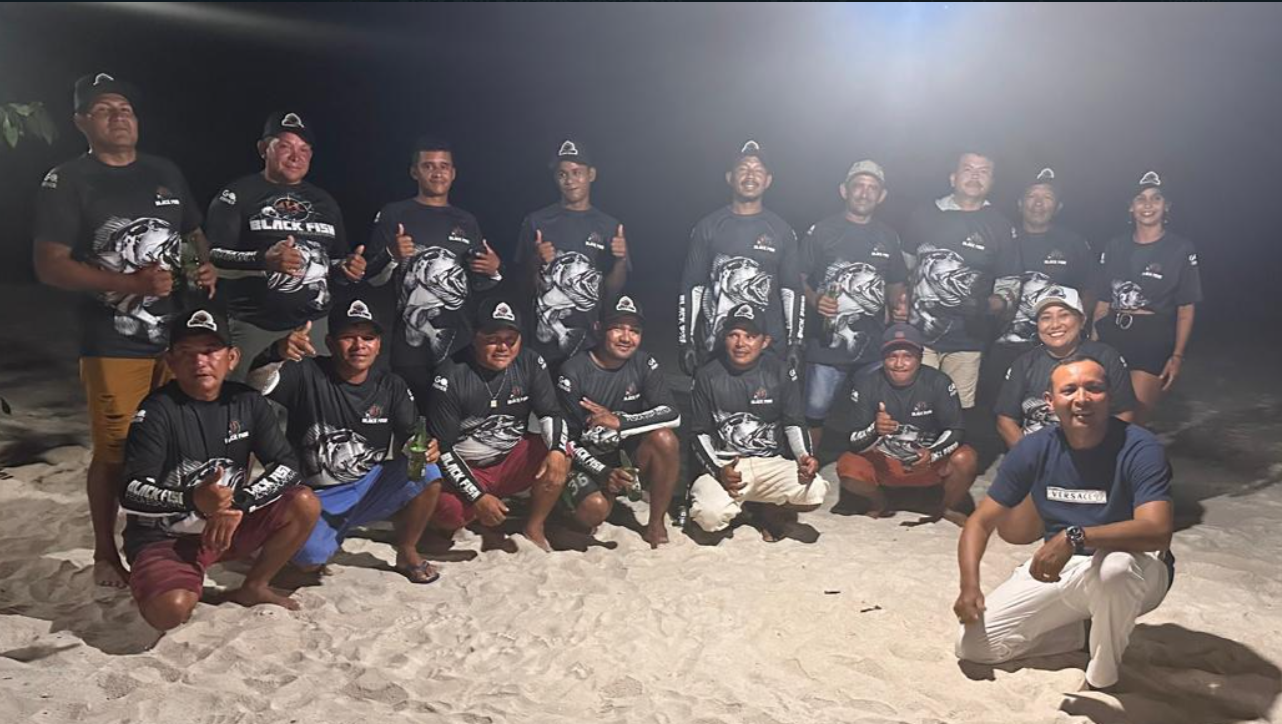 Group of people on a beach at night, wearing matching black sports jerseys with a fish graphic, posing for a photo with some giving thumbs up.