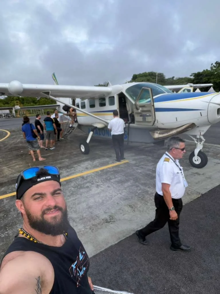 A man taking a selfie on a small airport tarmac with a small aircraft behind him. Several people are boarding the plane via a portable staircase, and a pilot or crew member is walking away from the plane. The sky is cloudy.