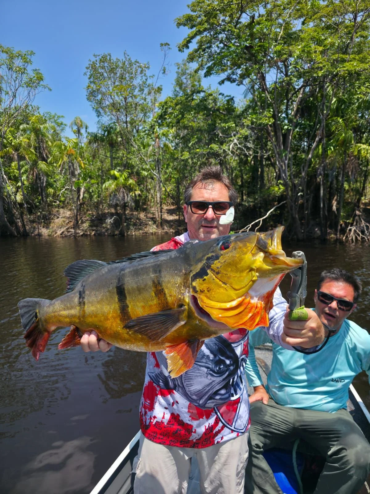 Man wearing sunglasses holding a large yellow and black fish with red eyes on a boat in a river, with another man sitting next to him, during a sunny day in a lush green forest.