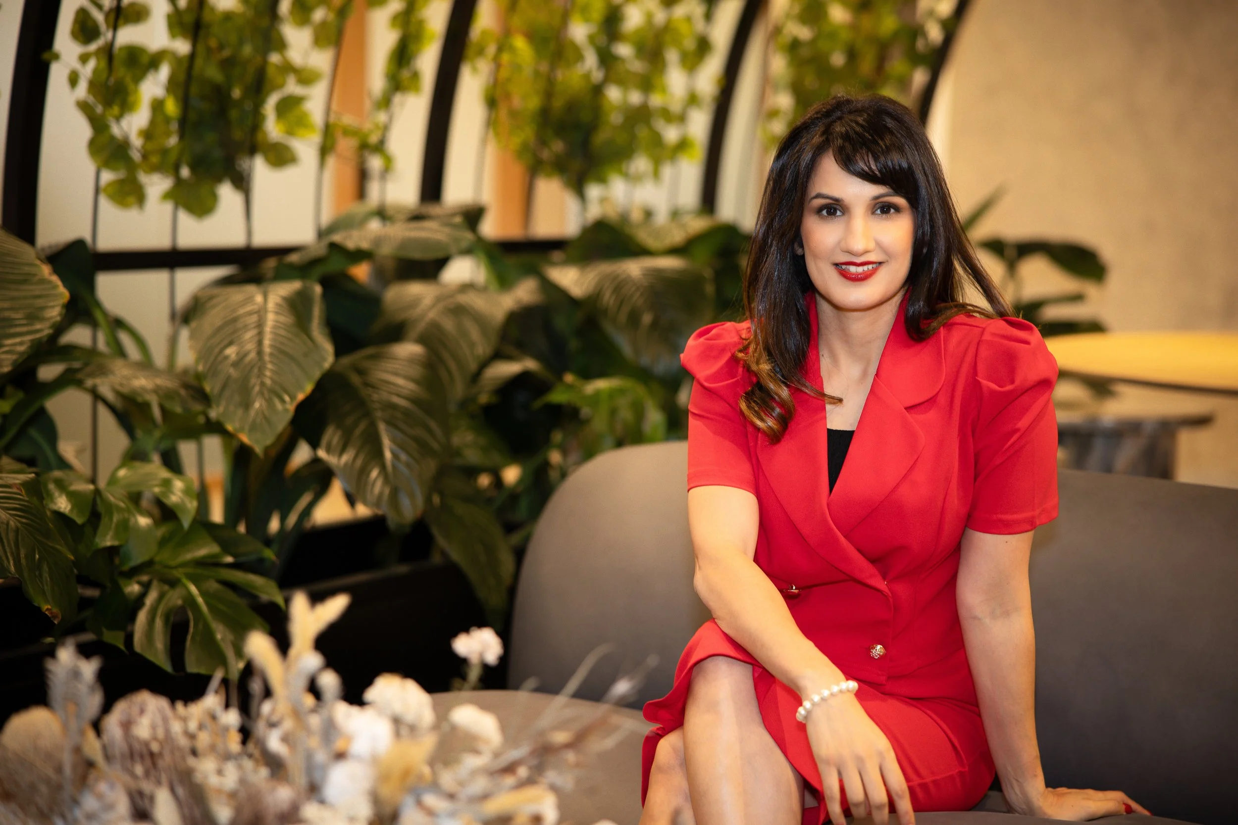 A woman in a red dress sitting on a sofa in a room with green plants, smiling at the camera.
