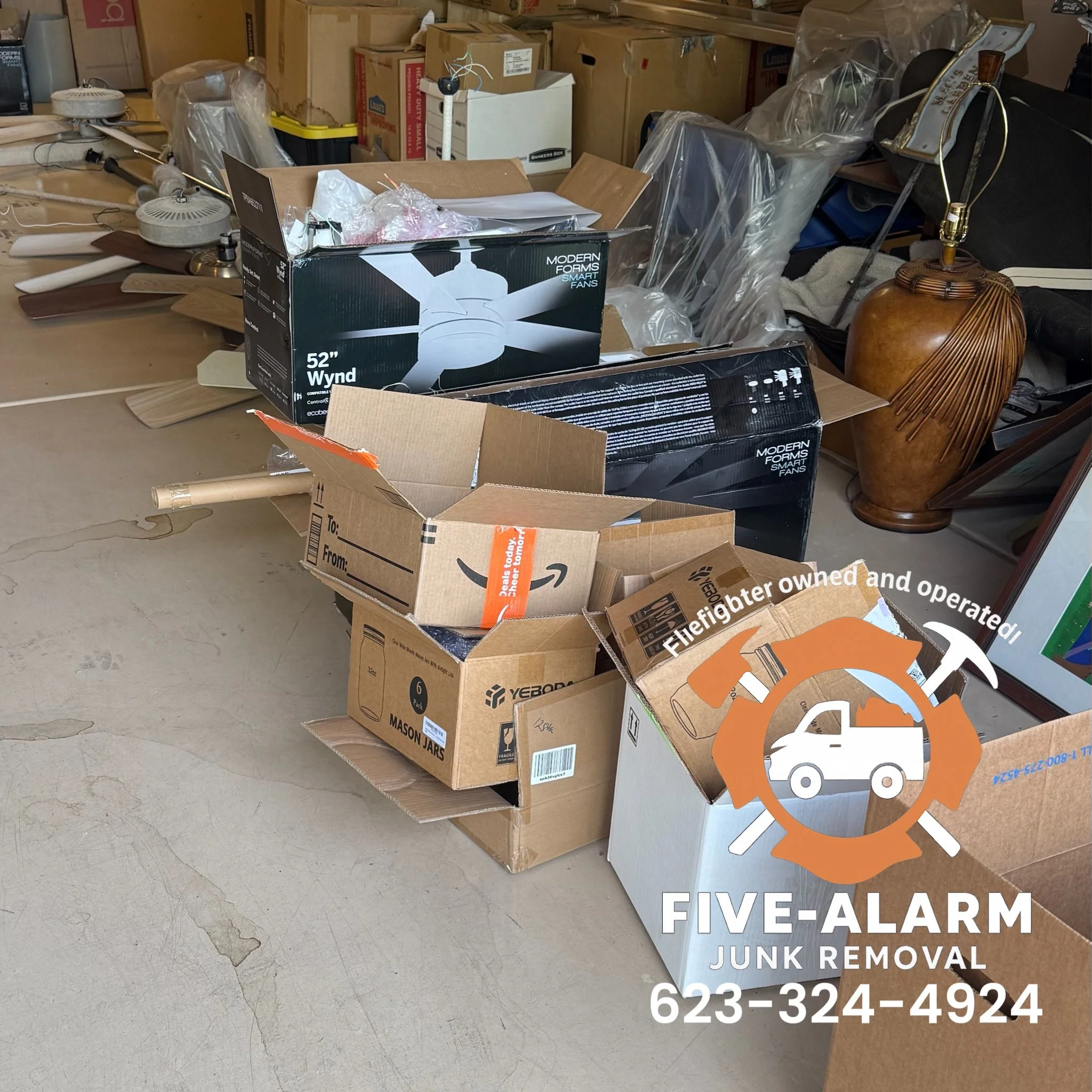 A cluttered workspace with cardboard boxes, some open and some closed, containing various items like a fan box, lamp base, and miscellaneous objects. In the background, there are plastic-wrapped items, a large decorative lamp, and some furniture. The
