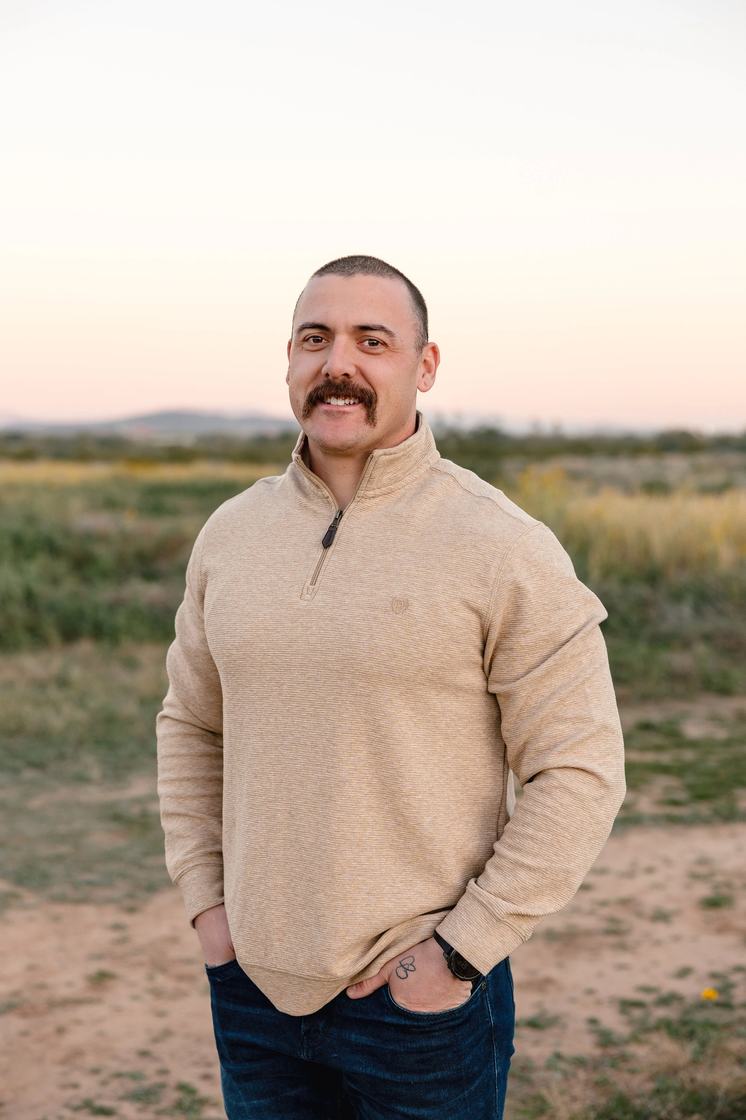 A man with a mustache and short hair standing outdoors at sunset, wearing a beige quarter-zip sweater and jeans, with one hand in his pocket.