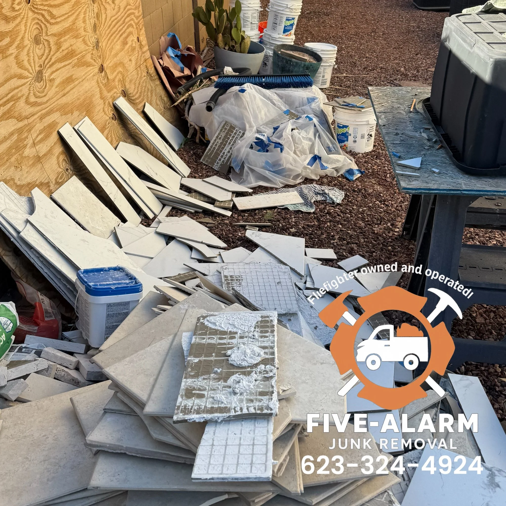 Piles of broken white tiles, construction debris, white buckets, and tools outside near wooden wall with a cactus plant.