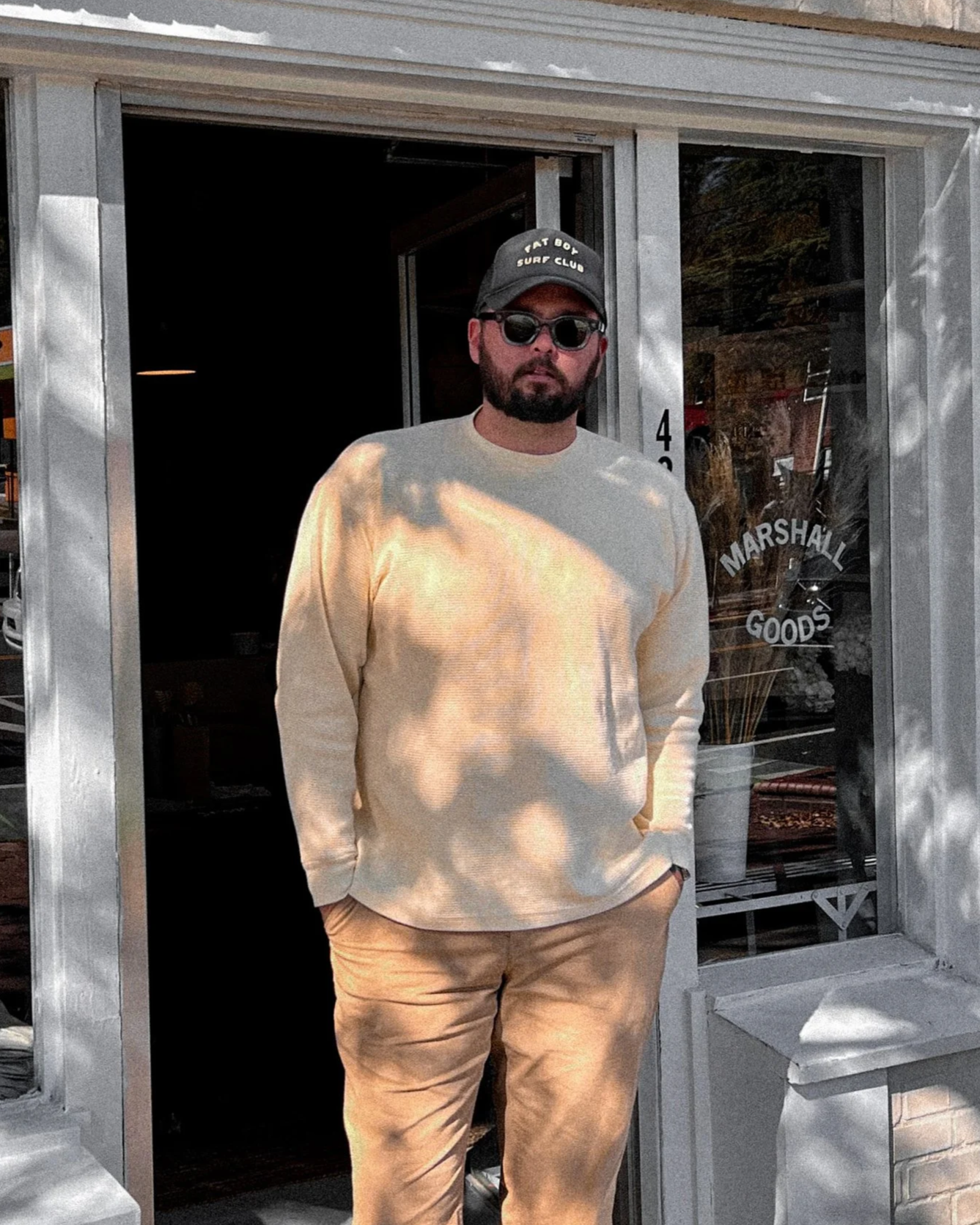 A man standing outside a store named Marshall Goods, wearing sunglasses, a black cap, a beige sweatshirt, and beige pants, with hands in pockets and shadow patterns on his clothing.